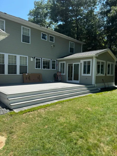 Back of a two-story house with a deck and enclosed sunroom, on a grassy lawn under a sunny sky.