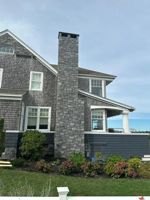 Stone house exterior with tall chimney, gray shingle siding, flower beds, and ocean view under a blue sky