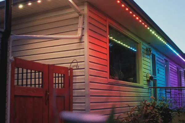 House exterior featuring permanent lighting along the roofline and window, casting colorful hues across the siding and a red gate. The lights create a modern, festive ambiance.