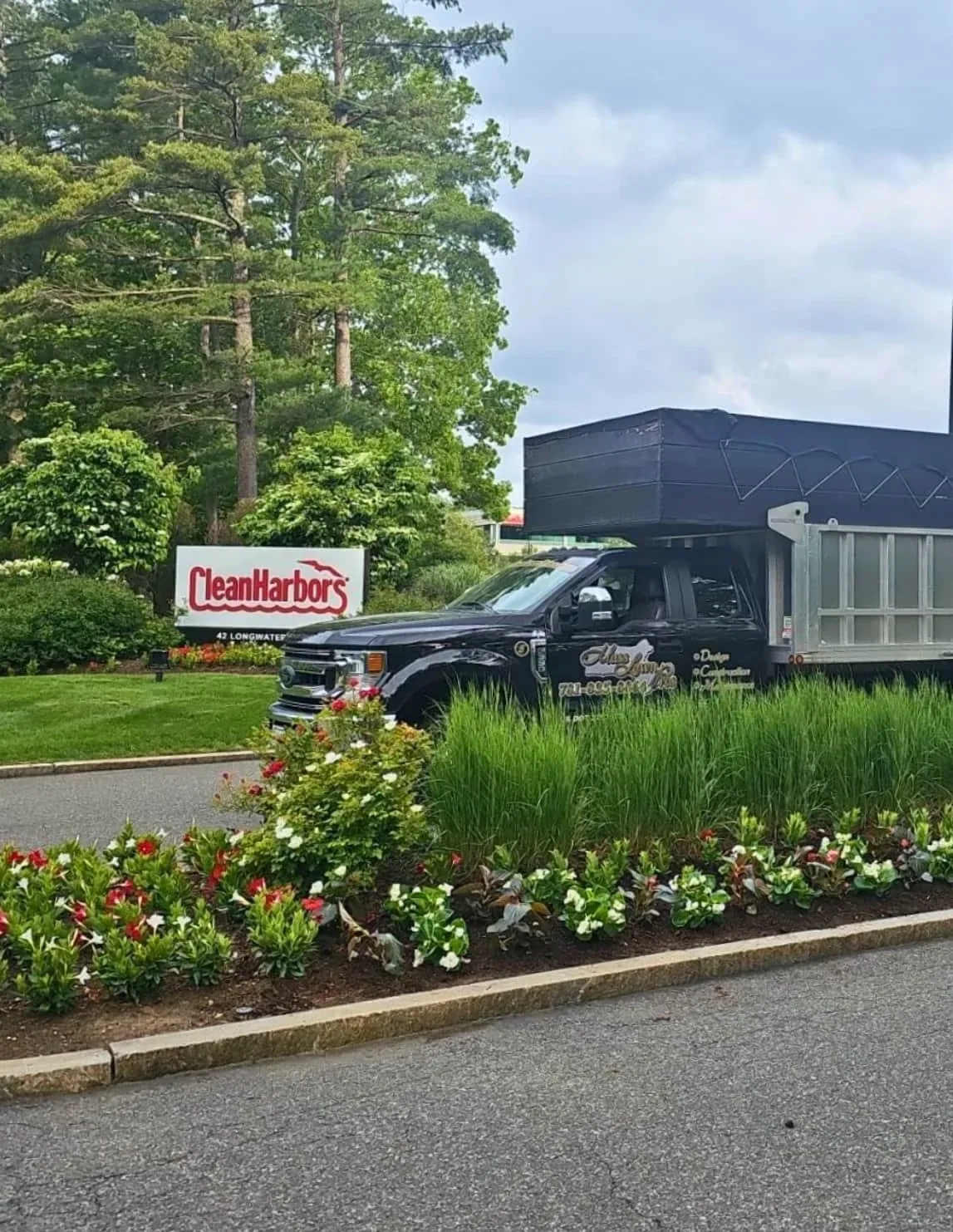 A black utility truck parked on a driveway next to a landscaped bed with colorful flowers and green grass, with trees in the background and a sign that reads 'CleanHarbors'.