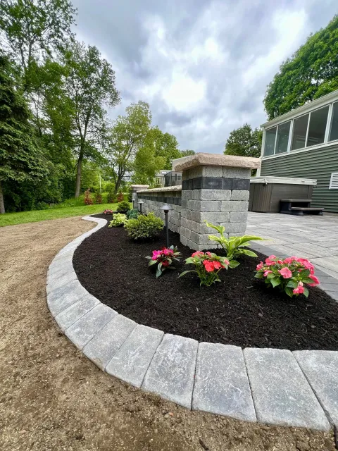 A landscaped backyard with a curved stone border, pink flowers, small shrubs, and a brick and stone wall. There is a patio with gray flooring and a gray building with enclosed porch. Trees and cloudy sky in the background.
