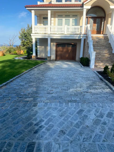 A driveway made of gray cobblestones leading to a two-story house with a garage and a staircase, surrounded by green lawn and landscaping, under a clear blue sky.