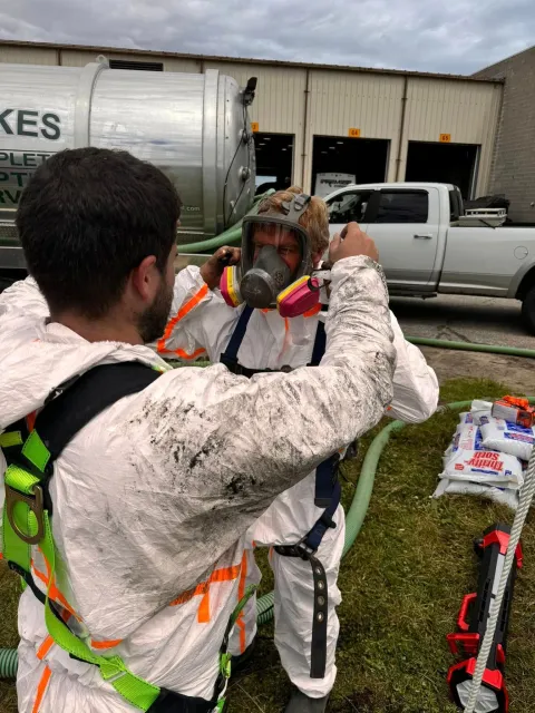 Two people in hazmat suits; one helping the other with their mask outdoors, next to a tanker truck.