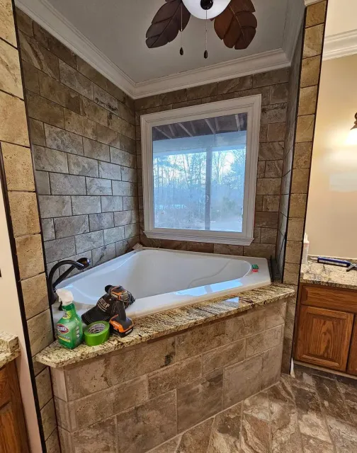 Corner bathtub in a tan stone-tiled bathroom beneath a window, with toiletries on the ledge.