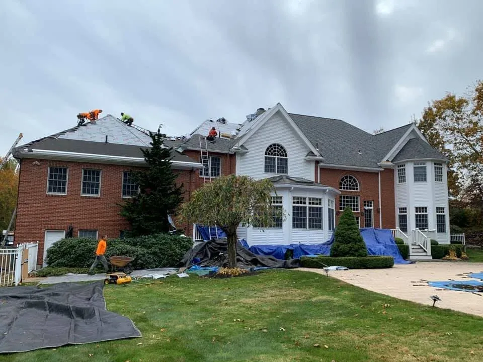 Construction workers repairing the roof of a large brick house that has sections of the roof covered with blue tarps, with construction equipment and materials on the lawn in front.