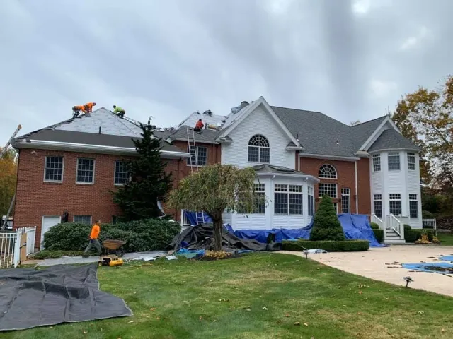 Construction workers repairing the roof of a large brick house that has sections of the roof covered with blue tarps, with construction equipment and materials on the lawn in front.