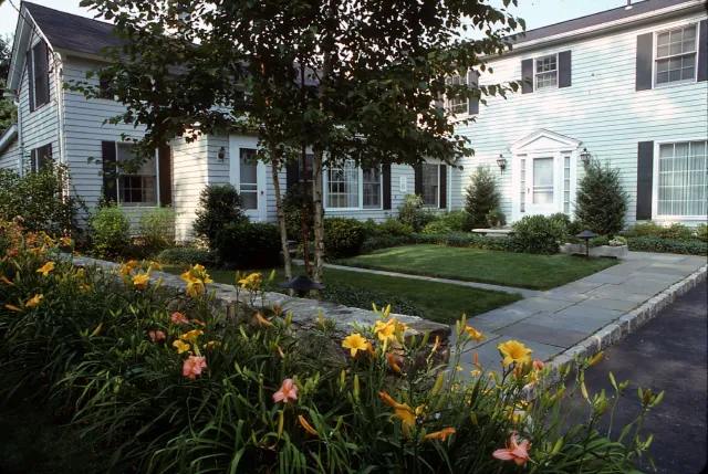 Front yard of a white house with black shutters, a well-maintained lawn, a sidewalk, and flowerbeds with yellow and pink flowers, and shrubs.