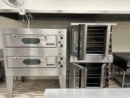 Commercial kitchen with two stainless steel ovens and a proofing rack on a tiled floor.