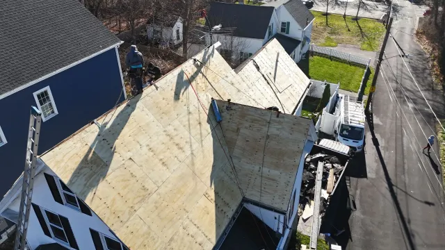 An aerial view shows workers installing new plywood roof decking on a home, with a construction van parked nearby.