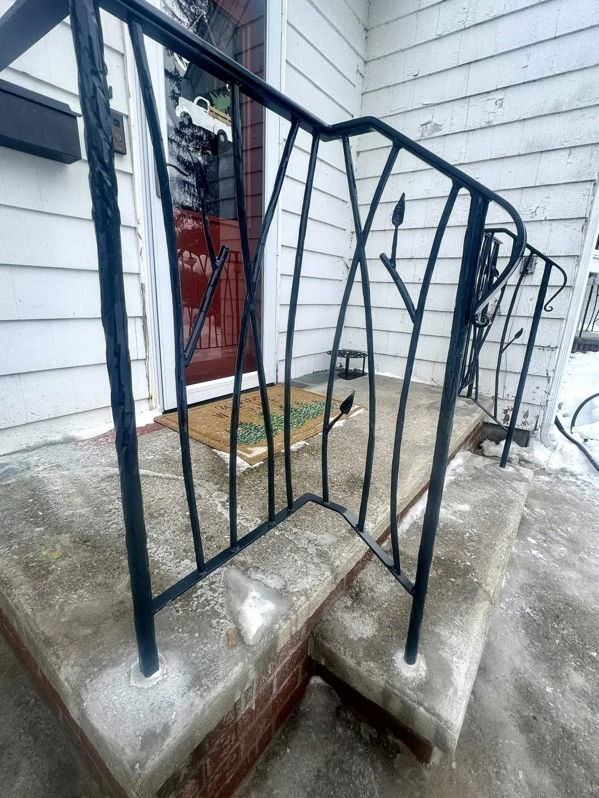 Black metal railing on concrete porch steps in front of a white house with siding. A red door with a decorative doormat is visible, along with snow on the ground.