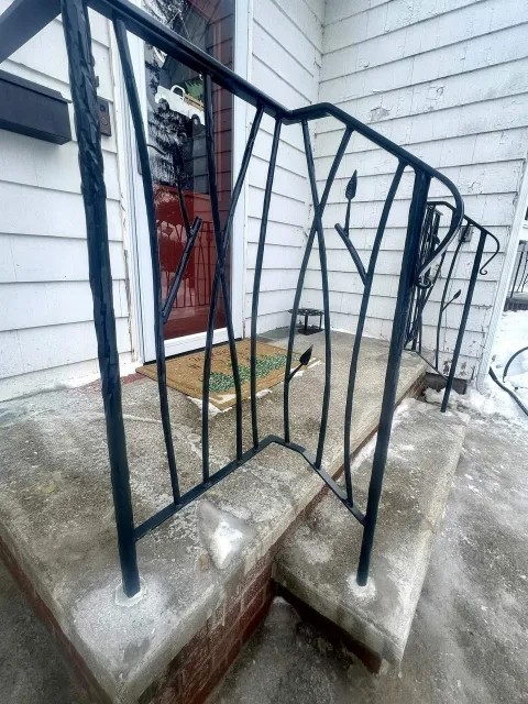 Black metal railing on concrete porch steps in front of a white house with siding. A red door with a decorative doormat is visible, along with snow on the ground.