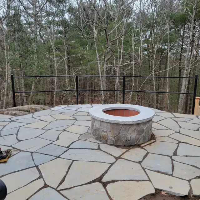 Stone patio with fire pit and black railing overlooking a forest.