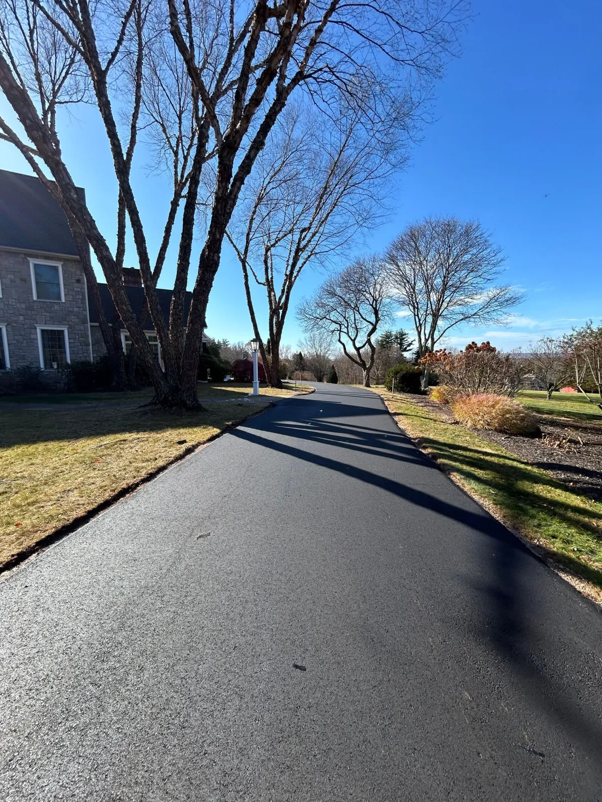 A freshly paved black asphalt driveway leads toward a stone house and bare trees under a clear blue sky.
