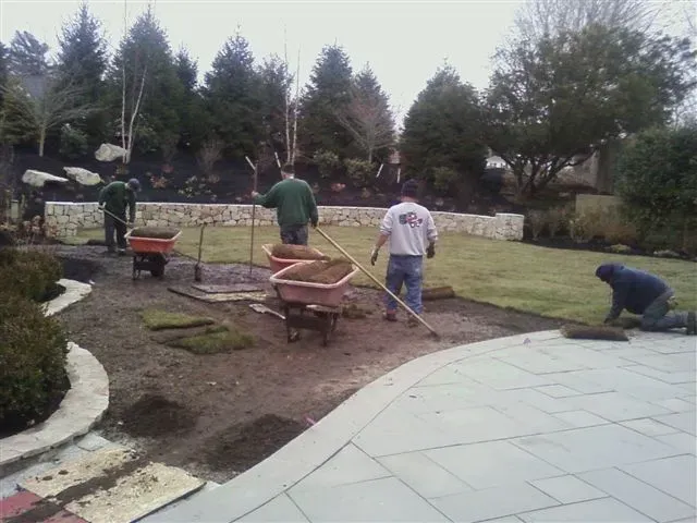 Workers shoveling and leveling soil in a landscaped garden beside a paved walkway