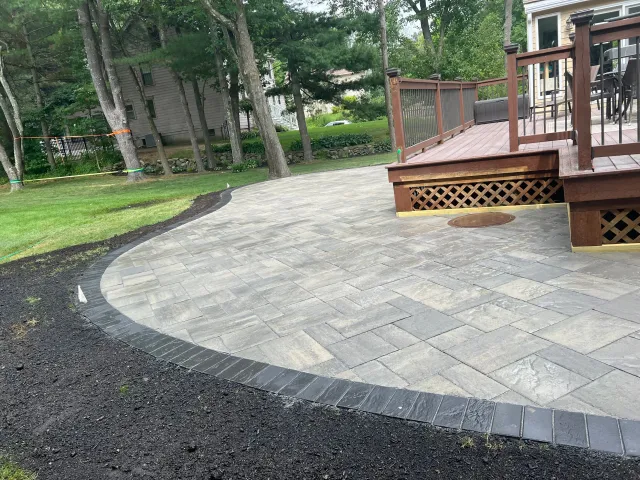 Newly constructed stone patio with a curved edge, surrounded by green grass and trees, and a wooden deck with outdoor furniture.