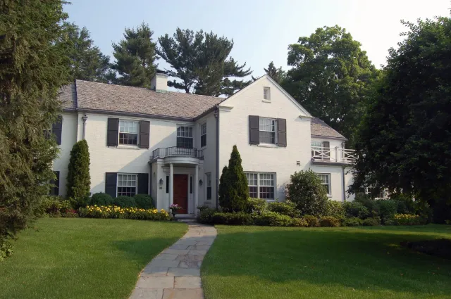 A two-story white house with black shutters, surrounded by a well-maintained lawn and garden, with tall trees in the background.