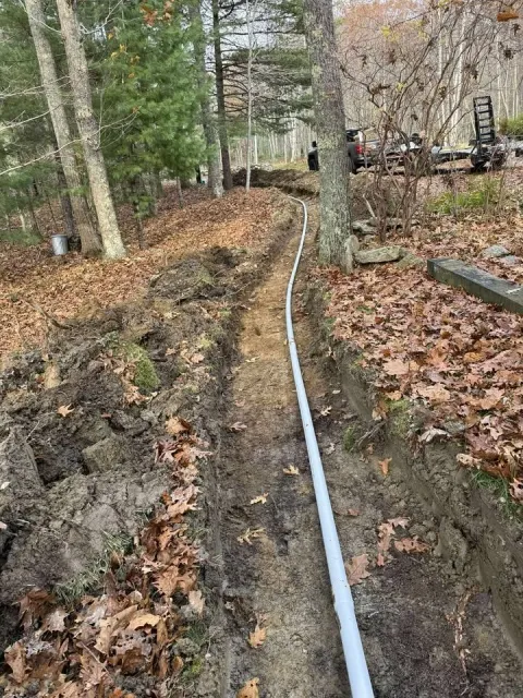 A narrow trench dug into the ground with a gray conduit pipe running through it, surrounded by fallen leaves and trees, in a wooded area near vehicles and outdoor equipment.