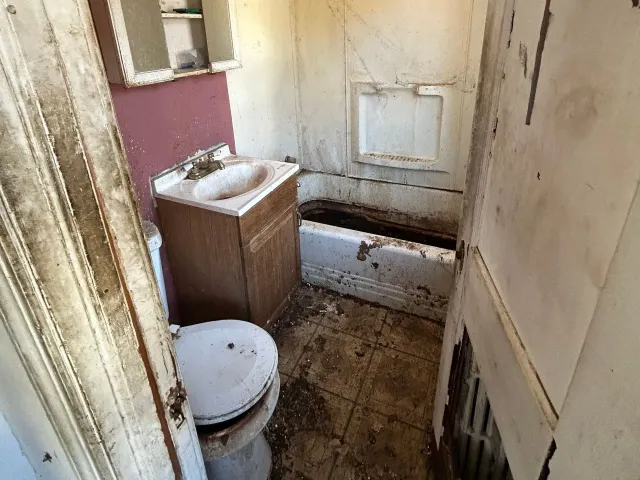 A dilapidated bathroom with a dirty toilet, sink, and bathtub. The walls are stained and peeling.