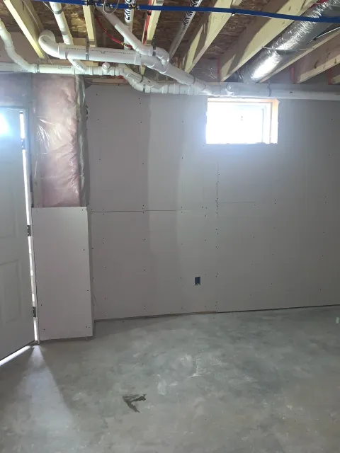 Interior view of a room under construction with drywall partially installed, a small window, and exposed ceiling with pipes and beams.
