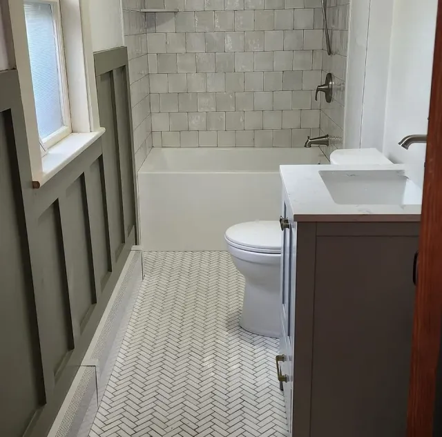 Small bathroom with a bathtub, toilet, and gray vanity with a sink. White tile wall behind the bathtub and a patterned tile floor.