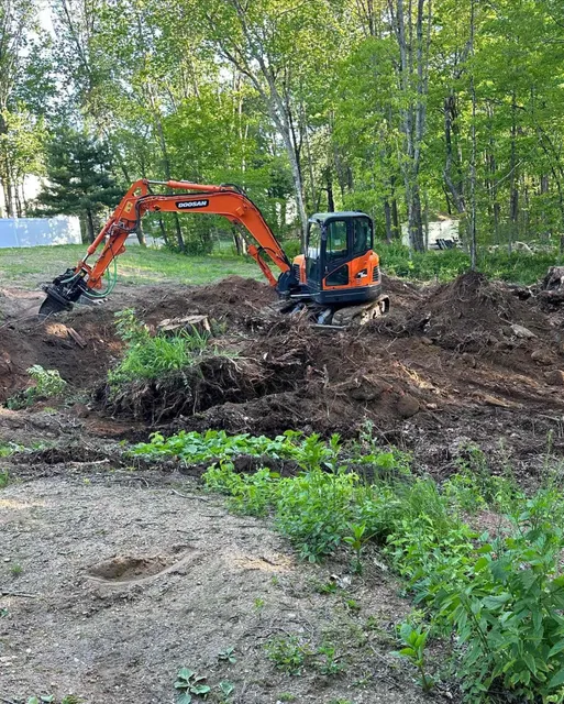 An orange excavator digging in a dirt area surrounded by green plants and trees.