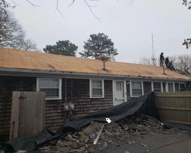 Roofers removing old shingles from a residential building on a cloudy day. Debris is covered by a tarp.