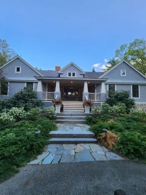 Gray house with a front porch, stone path, and lush greenery under a blue sky