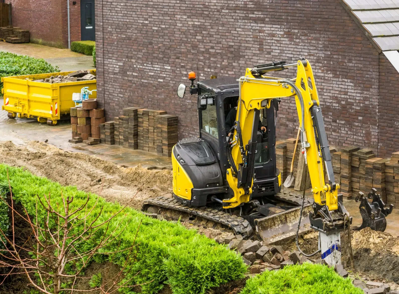 A yellow excavator is working on a construction site in front of a brick building.