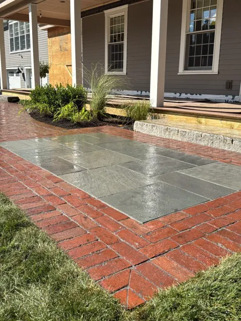 Wet stone patio bordered by red bricks leading to a porch with plants and a gray house exterior.