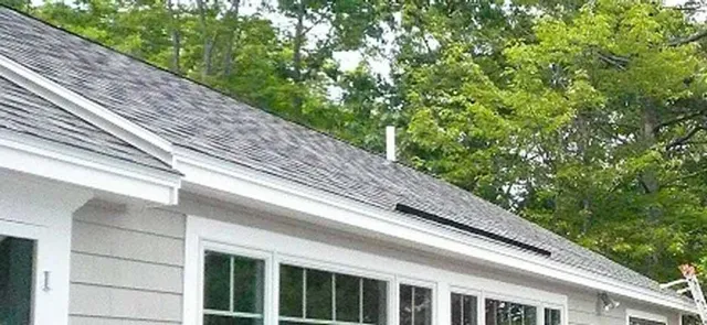 A gray-shingled house exterior with white trim, large windows, and a white vent pipe on a sloped roof near green trees.