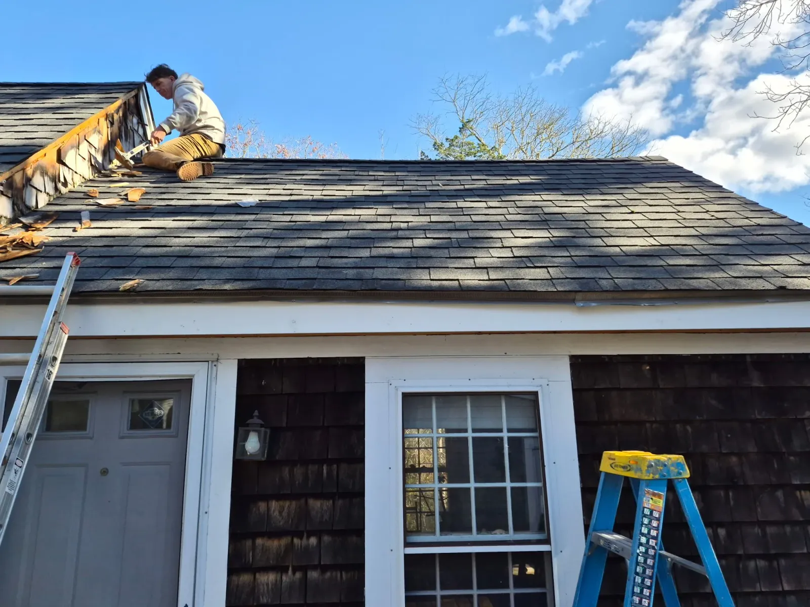 A person working on the roof of a house, sitting near the peak, with ladders on the ground. The house has a white door and window, with the roof covered in shingles. The sky is blue with some clouds.