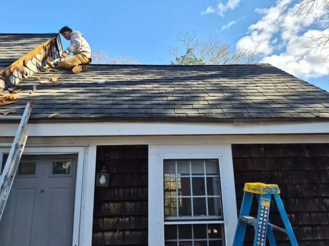 A person working on the roof of a house, sitting near the peak, with ladders on the ground. The house has a white door and window, with the roof covered in shingles. The sky is blue with some clouds.