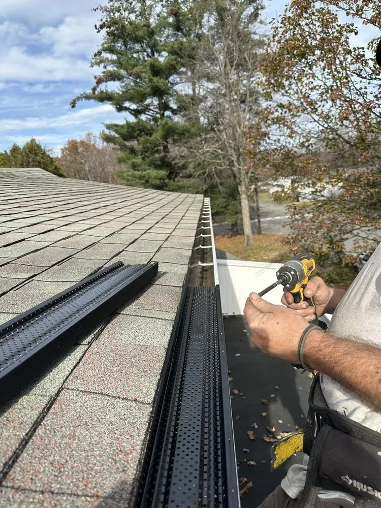 Worker installing black gutter guards on a shingled roof beside trees and a driveway.