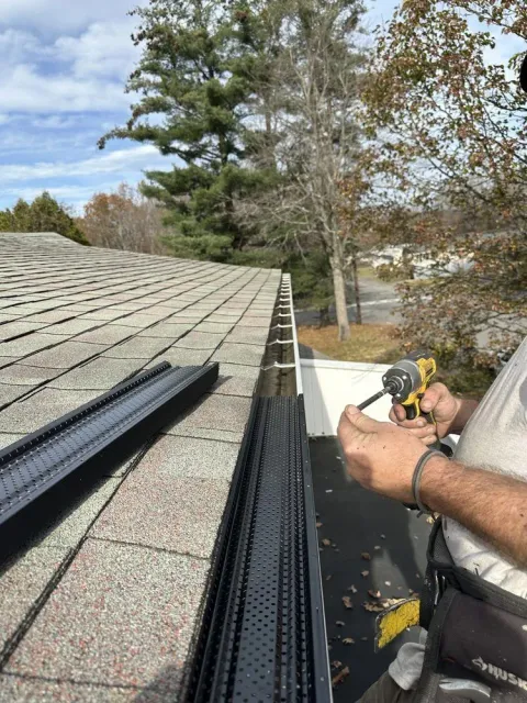 Worker installing black gutter guards on a shingled roof beside trees and a driveway.