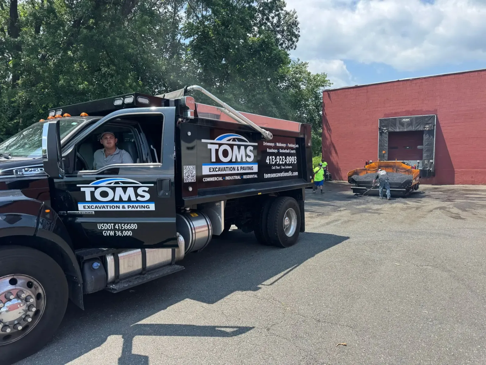 A black Tom's Dump Truck parked near a red brick building; a person in the driver's seat looks at the camera.