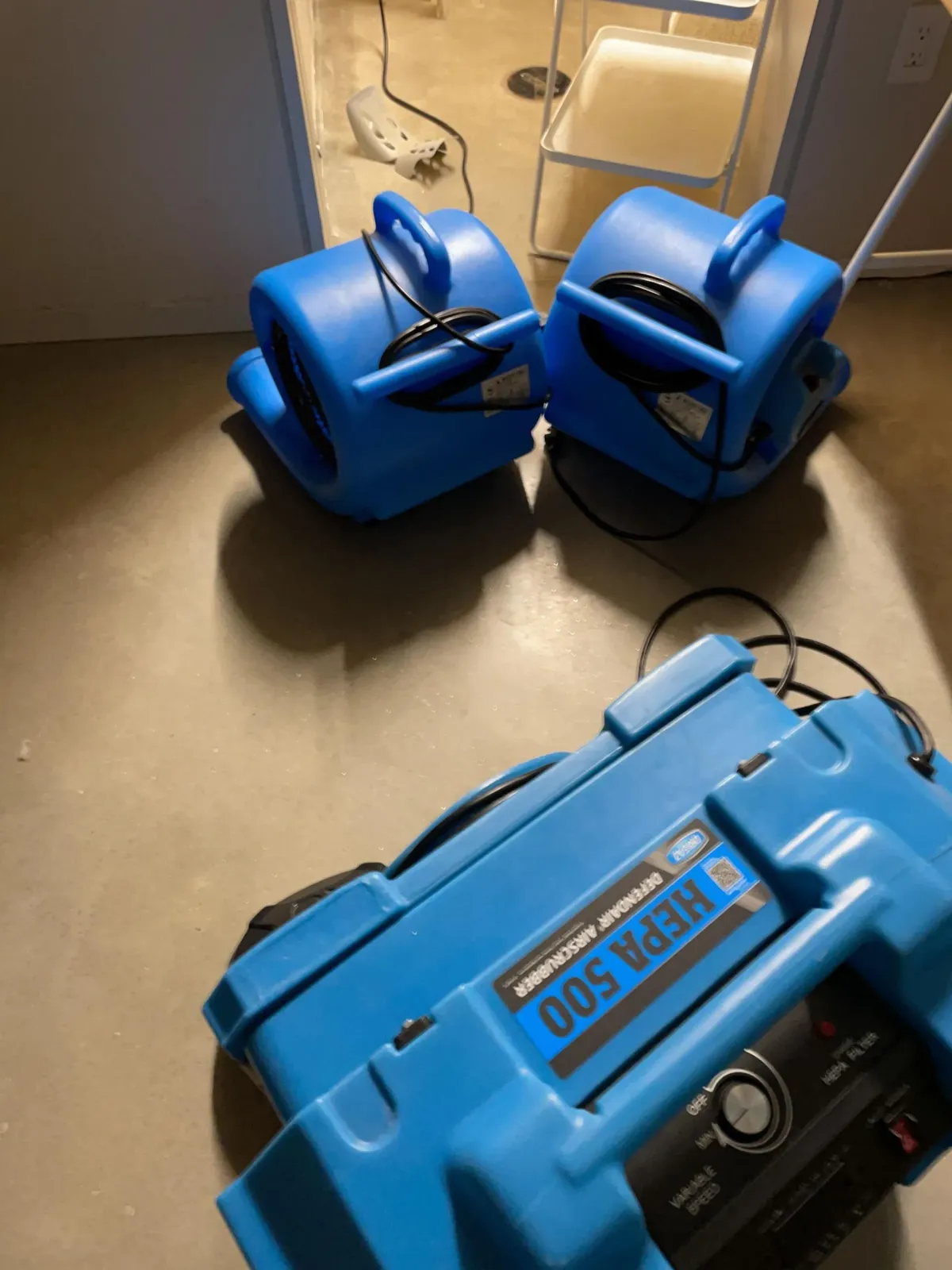 Blue battery charger with two blue battery packs on a floor near an outlet.
