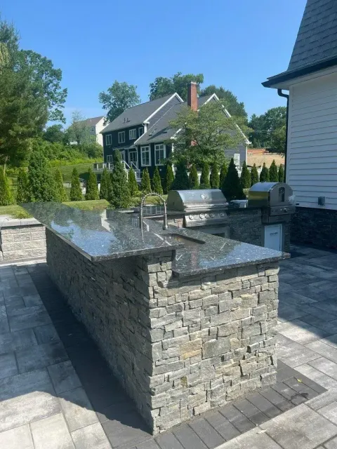 Stone outdoor kitchen island in a landscaped backyard beside a house, with trees and hedges in the background