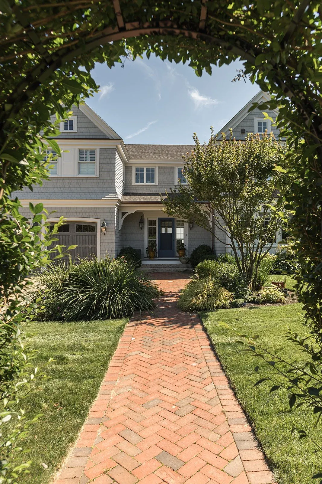 Brick walkway leading to a gray house with a front porch, surrounded by green grass, bushes, and trees, seen through an arched hedge.