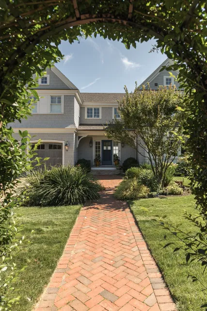 Brick walkway leading to a gray house with a front porch, surrounded by green grass, bushes, and trees, seen through an arched hedge.