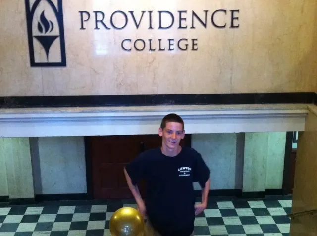 Person smiling in front of Providence College sign, wearing a dark t-shirt, standing on a black and white tiled floor.