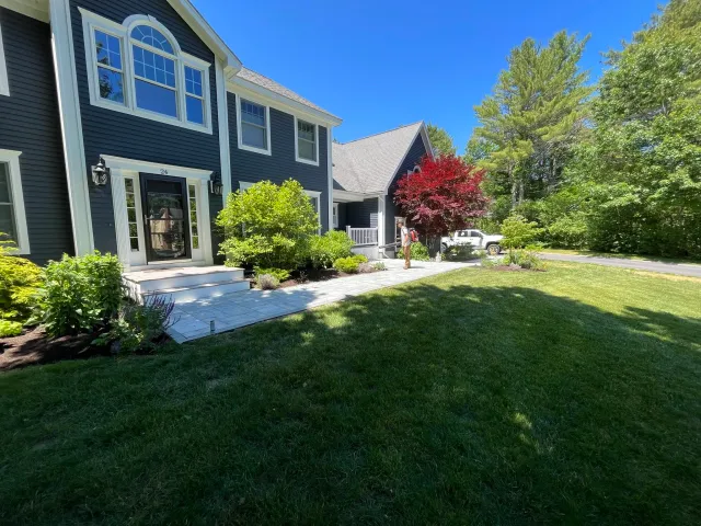 A blue house with white trim and a black front door, surrounded by a well-maintained yard with green grass, shrubs, and trees. A person wearing a hat and carrying a backpack is walking on a paved pathway.