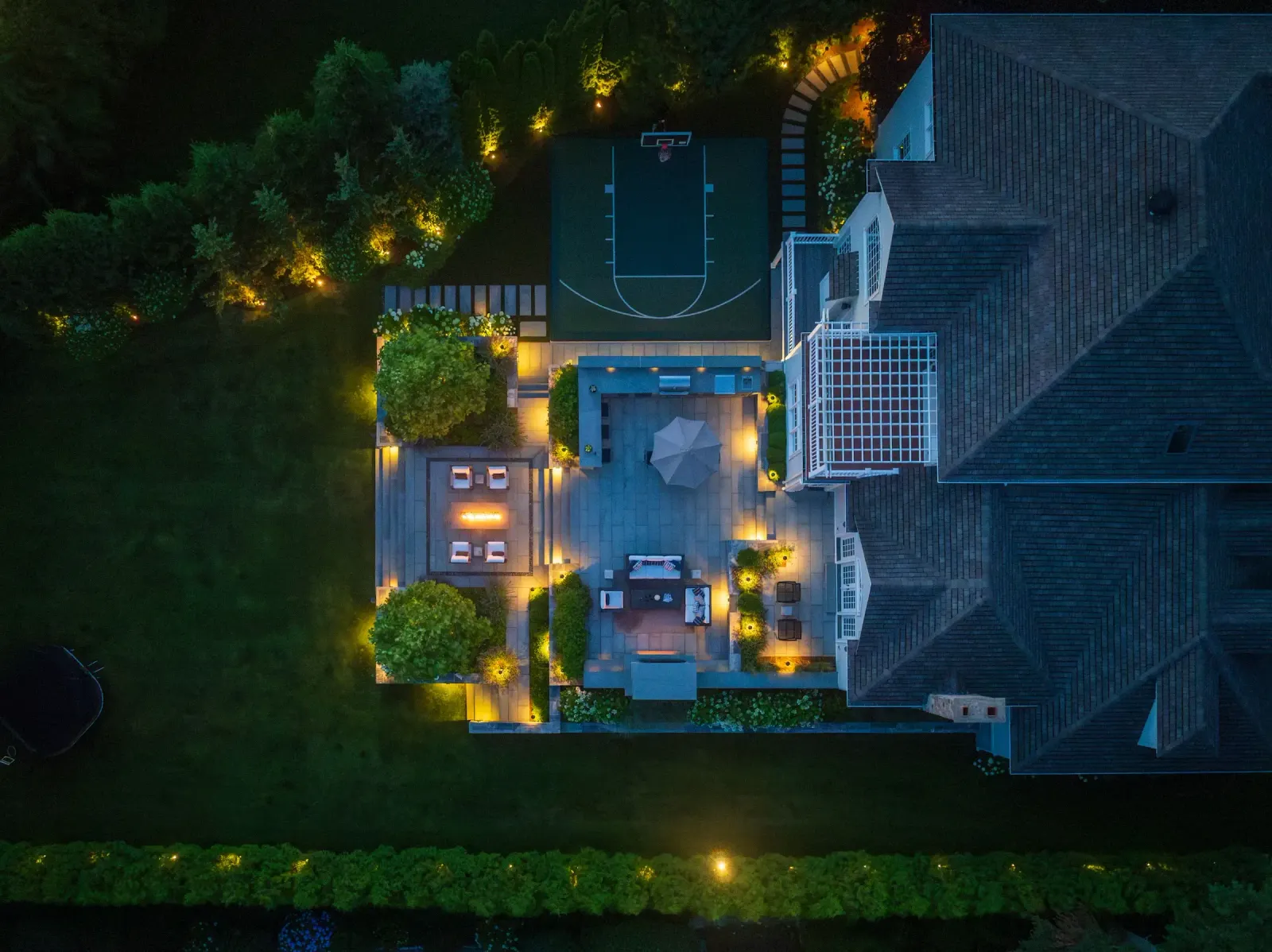 An aerial bird's-eye view of a Sudbury, MA estate at twilight. Warm landscape lighting illuminates a multi-level stone patio with a lounge area, outdoor kitchen, and fire pit. Next to the patio is a custom dark green basketball half-court, all surrounded by a manicured lawn and privacy hedges.