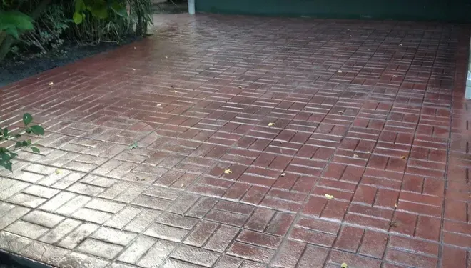 A wet, reddish-brown driveway featuring a herringbone brick pattern, with a dark green garage door in the background.
