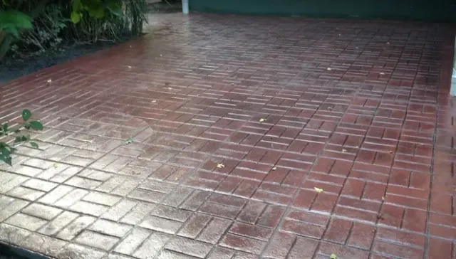 A wet, reddish-brown driveway featuring a herringbone brick pattern, with a dark green garage door in the background.