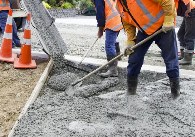 Workers in orange vests pouring wet concrete on a road beside traffic cones.