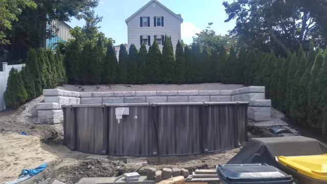 Backyard with above-ground pool construction, retaining wall, evergreen trees, and a house in the background.