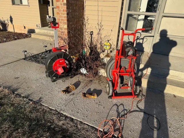 Outdoor scene with a concrete sidewalk next to a house wall. There is a red hose reel, a red pressure washer, and a person's shadow holding a camera. Equipment is placed near a small tree with dried branches, and the background includes a window and part of the house exterior.