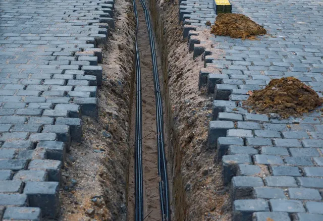 Open trench cuts through blue-gray brick pavement with exposed cables inside.