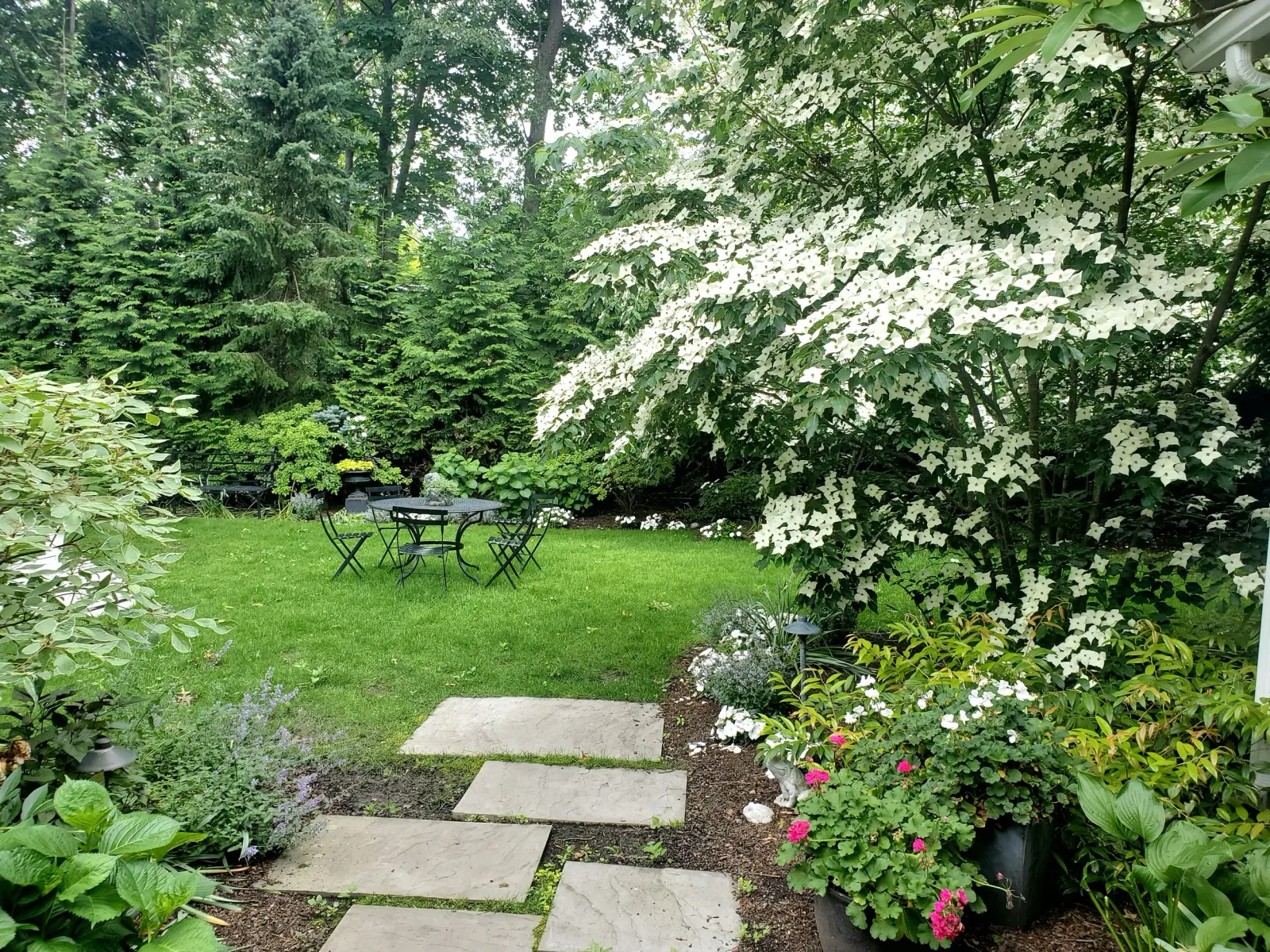 A lush backyard garden with a stone pathway, green grass, blooming white and pink flowers, trees, and outdoor black metal furniture.