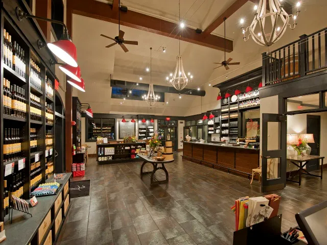 Interior of a wine shop or boutique with shelves filled with wine bottles, a checkerboard-patterned ceiling, chandeliers, ceiling fans, and a wooden counter for customer service.